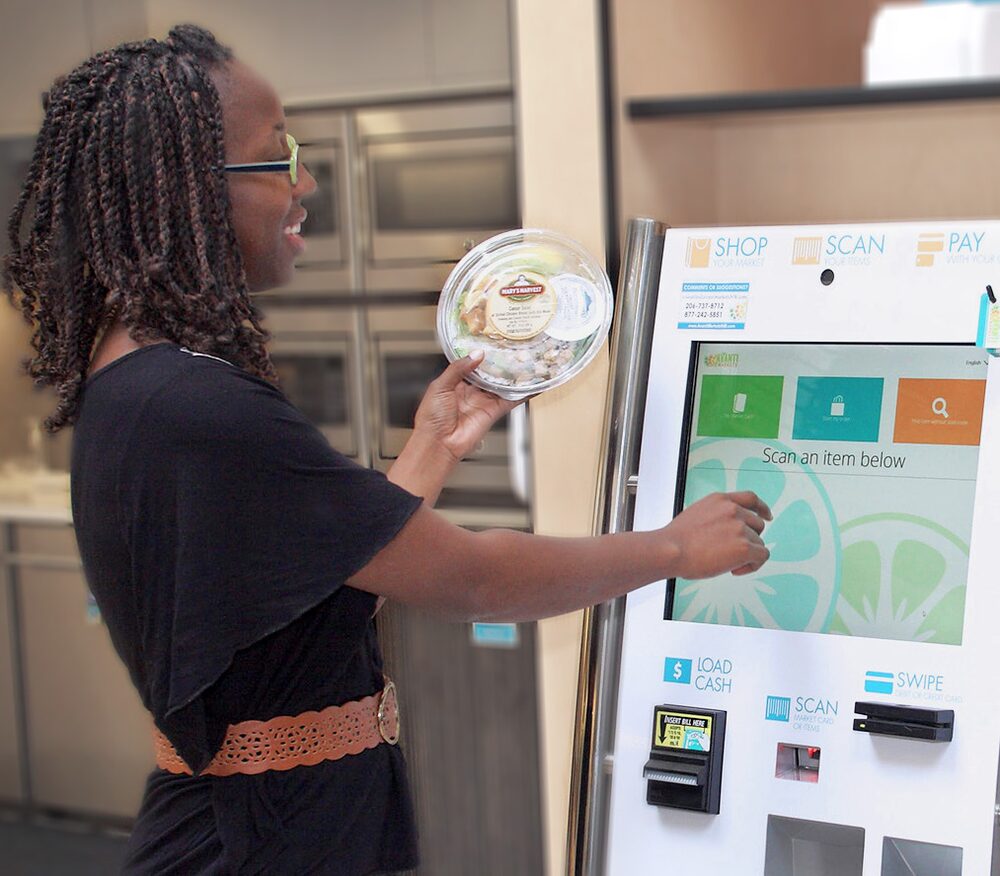 An employee holding a fresh salad bowl while scanning it at a micro-market self-checkout kiosk.