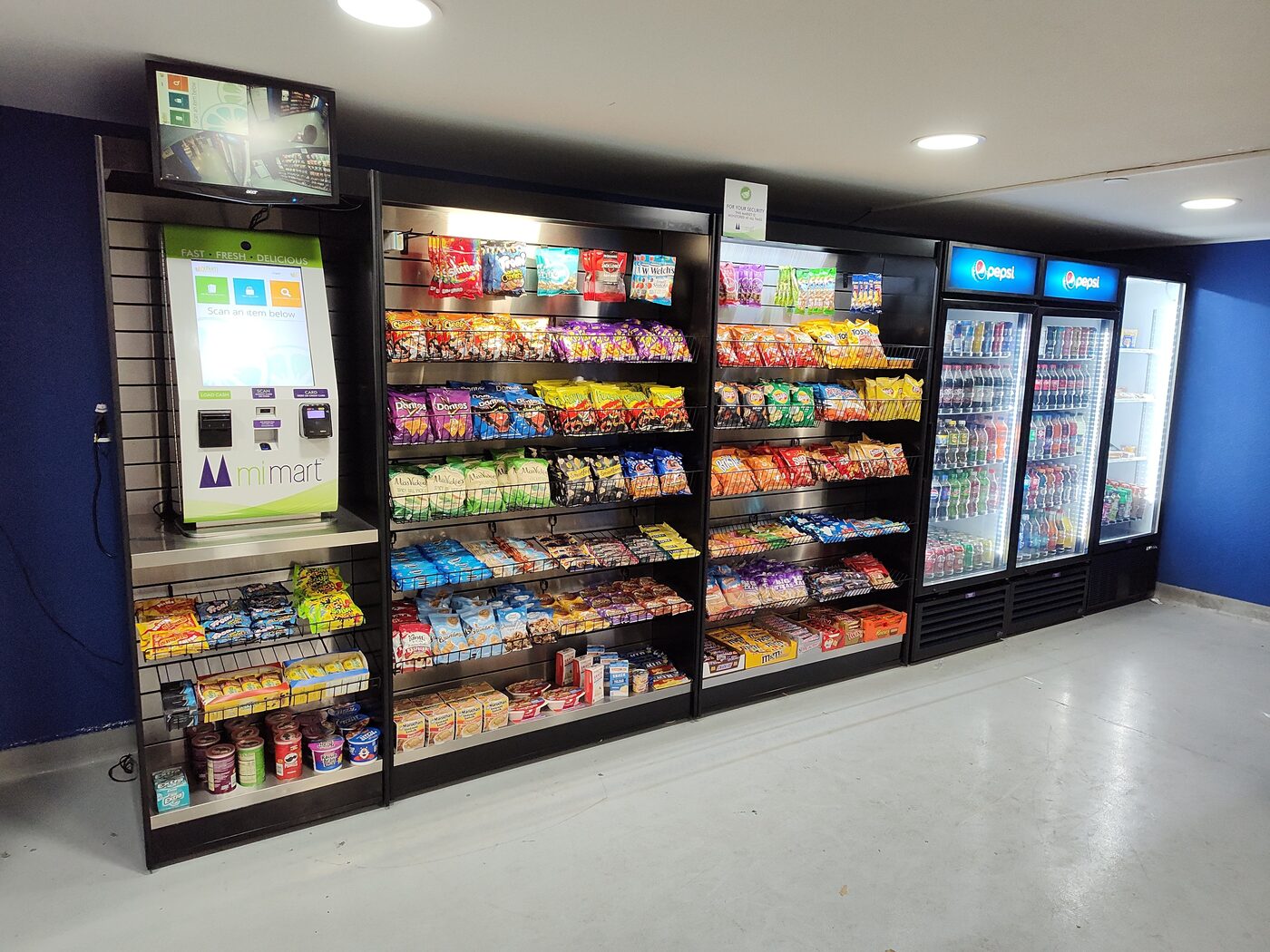 An installed MiMart micro-market: a self-checkout kiosk beside open shelving of snacks and three glass-door coolers of drinks.