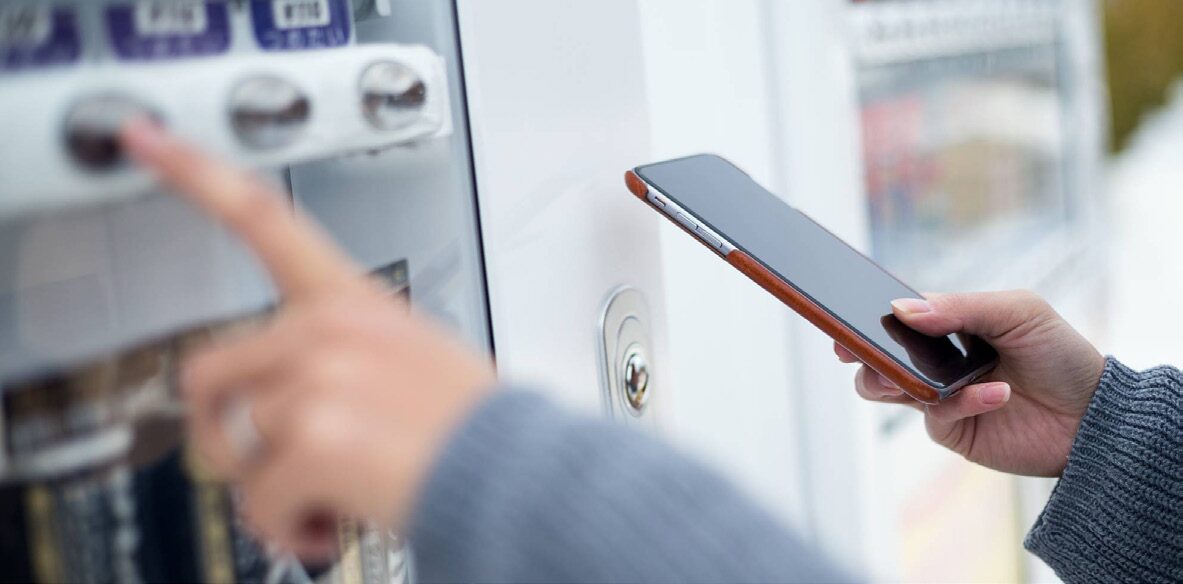 A person holding a phone up to the contactless reader on a vending machine while pressing a selection button.